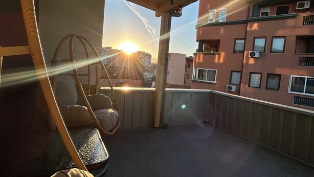 Un balcone con un'amaca sospesa, vista su edifici e un tramonto.
