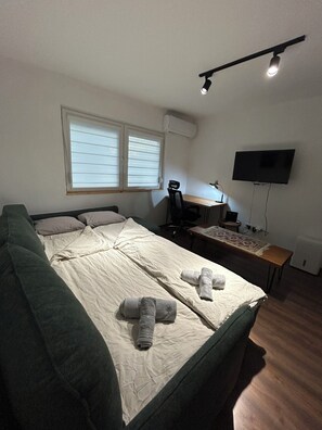 A bedroom with a bed, a desk, a chair, a television, and a window with blinds. - Affordable Cozy Apartment in Skopje (Skopje)