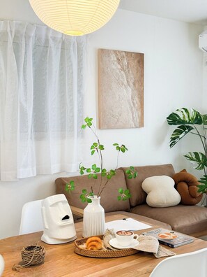 Ein gemütliches Wohnzimmer mit einer Couch, einem Couchtisch mit einem Croissant, einer weißen Vase mit Grünpflanzen und einem Gemälde an der Wand.