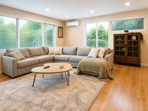 A living room with a sectional sofa, a round coffee table, a wooden cabinet, and large windows with blinds.