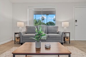 Ein Wohnzimmer mit grauem Sofa, zwei Beistelltischen, einem Holztisch, einem Blumentopf und Blick durch ein Fenster.