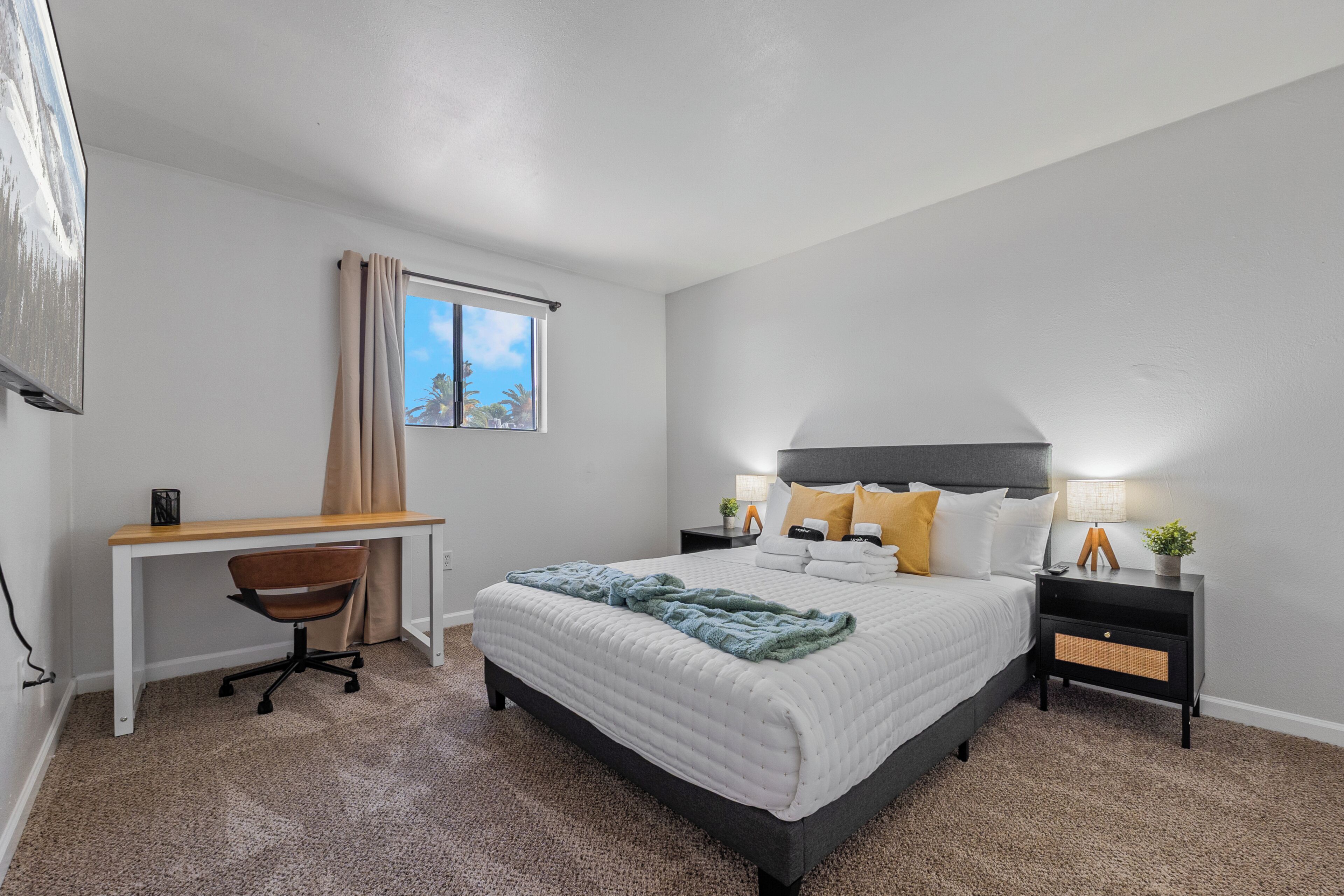 A bedroom with a bed, desk, chair, nightstands, and a window with curtains.