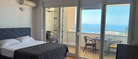 A bedroom with a bed, a small table, and a chair, a balcony with a view of the sea, and an air conditioning unit on the wall.
