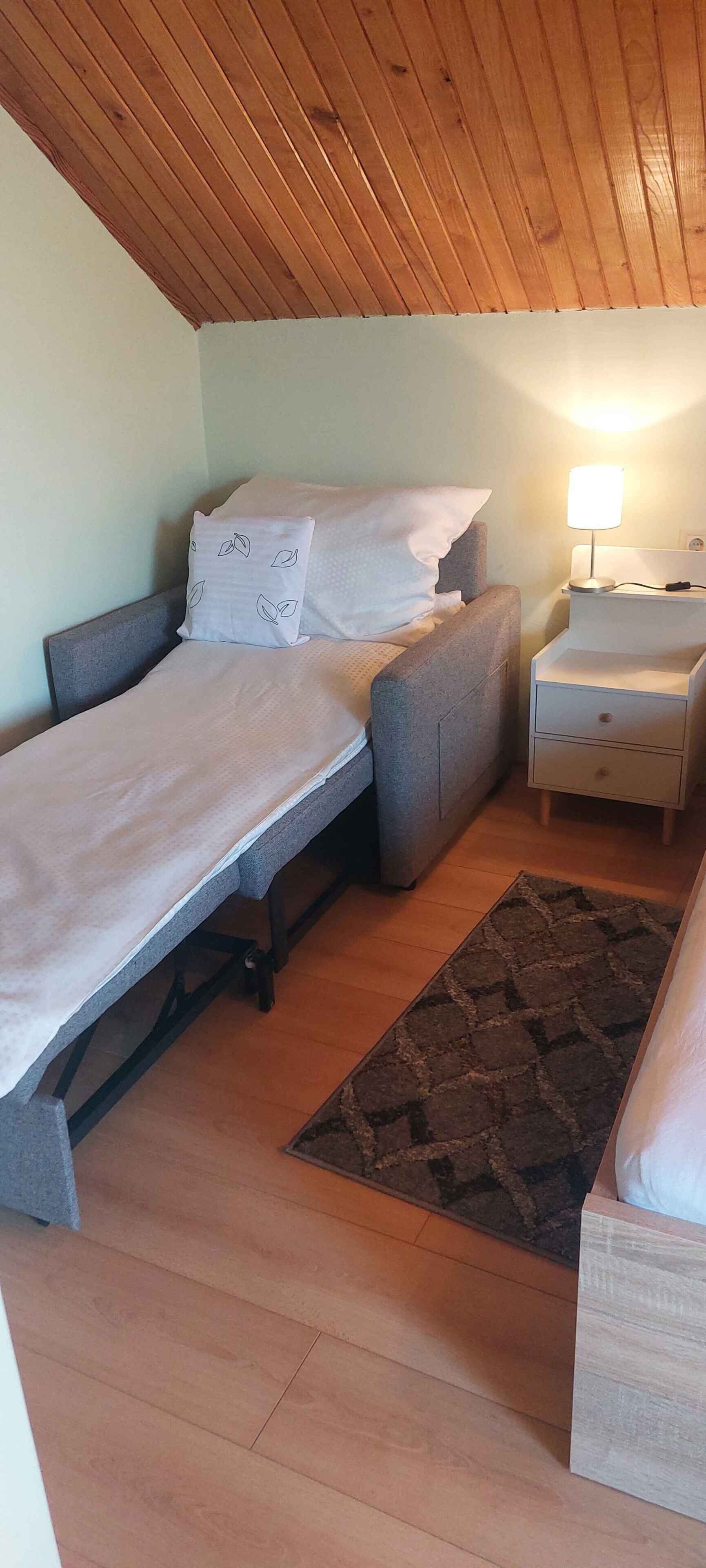 Een kleine, gezellige kamer met een bed, nachtkastje, lamp en houten plafond.