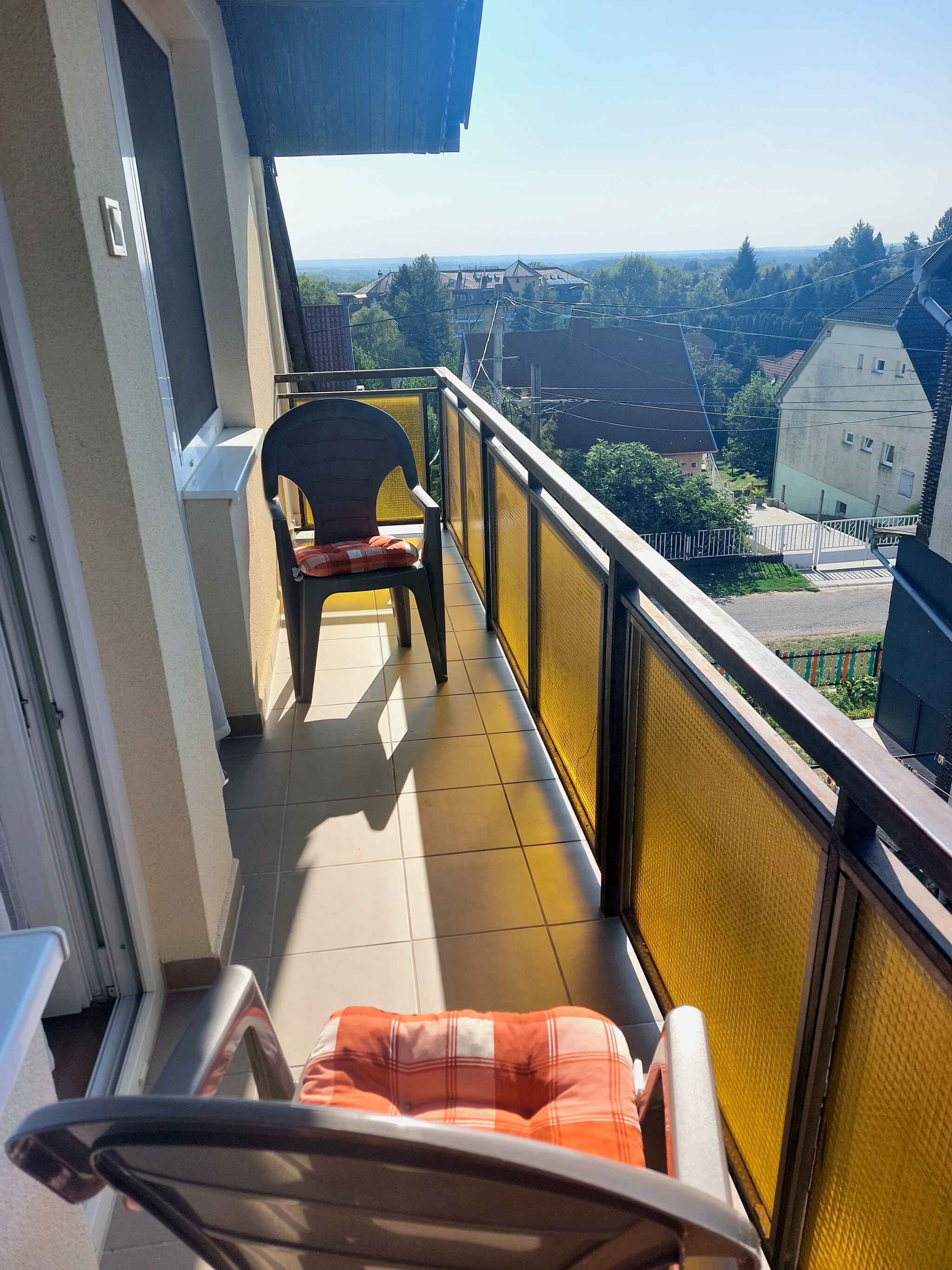 Un balcón con una silla y un cojín, vistas del vecindario y un edificio al fondo.