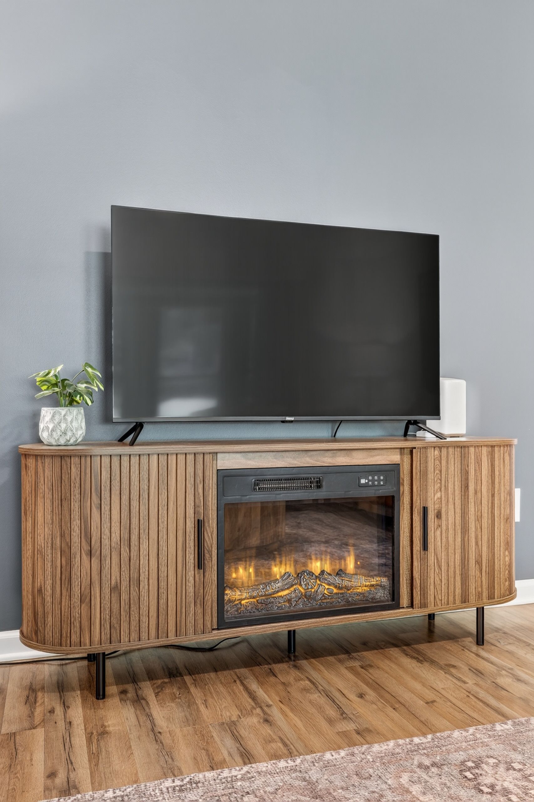 Un salon moderne avec un téléviseur à écran plat fixé sur un meuble en bois, une cheminée située sous le téléviseur et une petite plante posée sur le meuble.