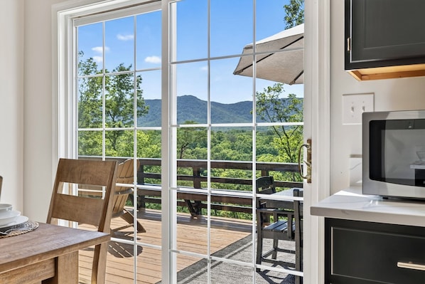 A room with a large window, a wooden deck, a table with chairs, and a television.