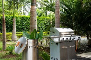 A stainless steel outdoor grill with a hood, a metal pot, and a control panel, set in a landscaped garden with palm trees and a hedge. - Luxury Villa in Santa Fe de Antioquia (Casa Sereno) (San Nicolas)