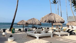 A beach with palm trees, thatched umbrellas, and sun loungers. - Beachfront Penthouse w Rooftop Pools Views (Punta Cana)
