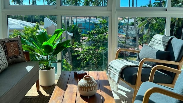 A room with a large window, a wooden table, a sofa, and a potted plant. - Beachfront Penthouse w Rooftop Pools Views (Punta Cana)