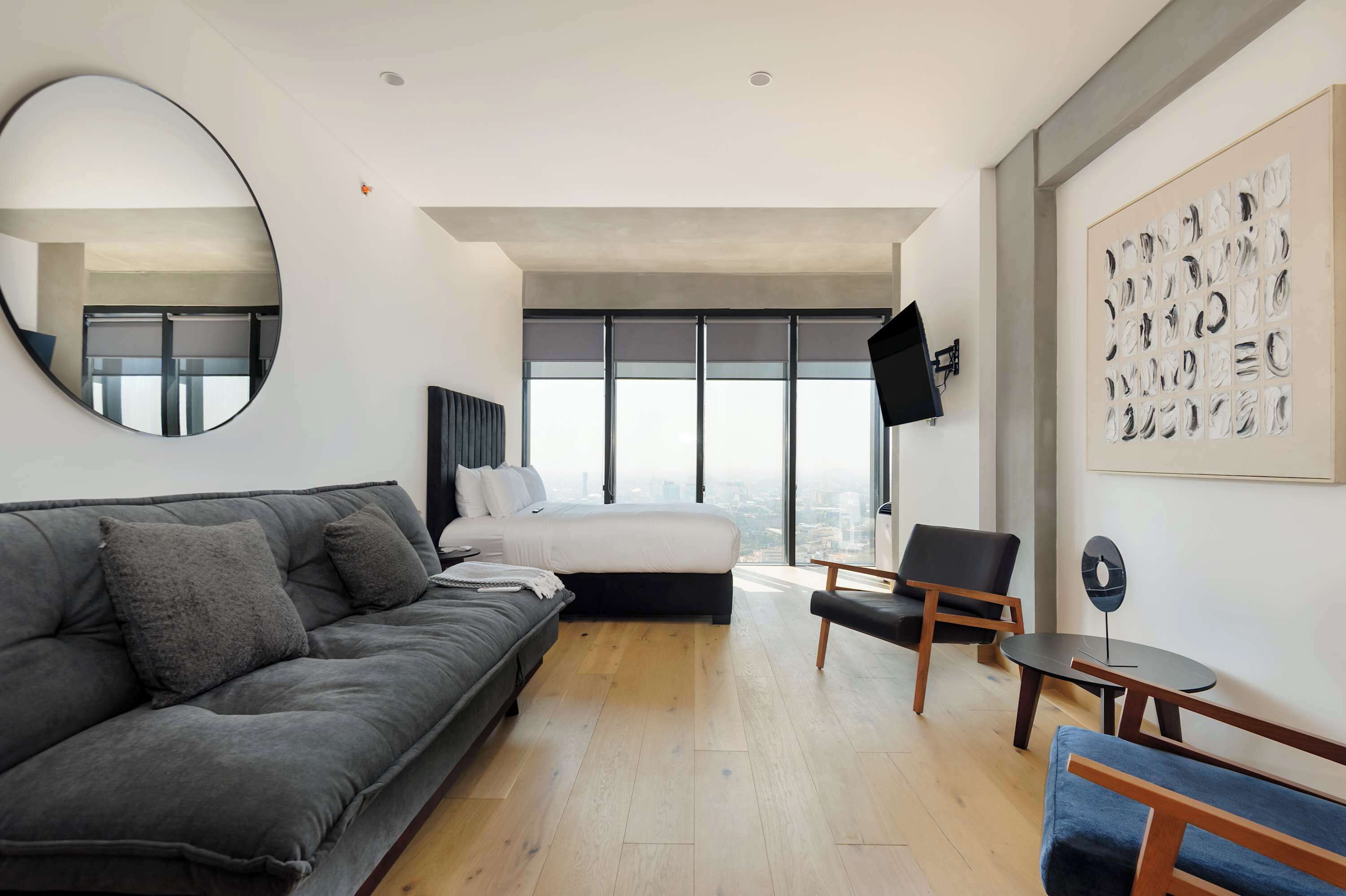 Una habitación de hotel moderna con una cama grande, un sofá gris, una mesita pequeña, una silla, un espejo redondo y vistas a la ciudad a través de amplias ventanas.