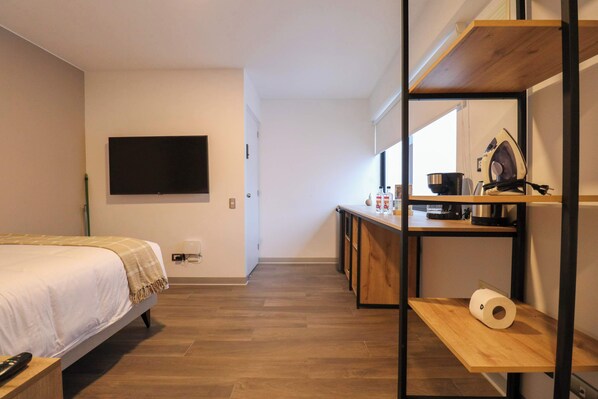 Ein Schlafzimmer mit einem Bett, einem Fernseher, einem Schreibtisch mit Wasserkocher und einem Holzregal mit Toilettenpapier.