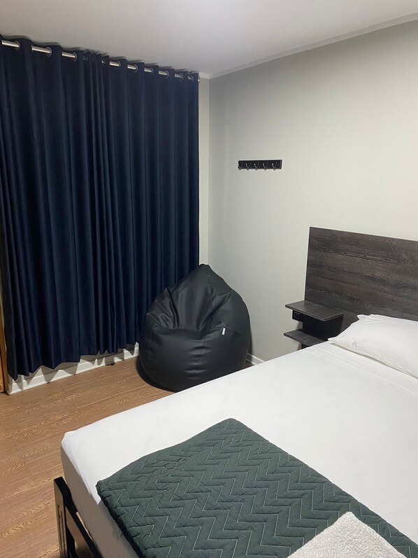 Una habitación de hotel con cama, un sillón tipo bean bag, una mesita de noche y una cortina oscura.