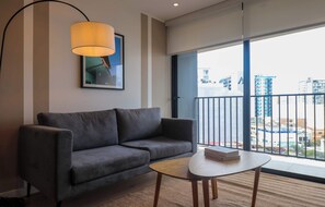Ein modernes Wohnzimmer mit einem grauen Sofa, einem kleinen runden Tisch, einer Lampe und einem großen Fenster mit Blick auf Gebäude.