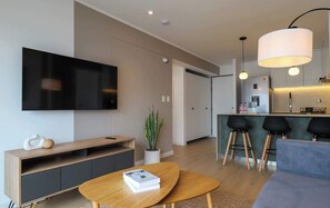 Ein modernes Wohnzimmer mit Flachbildfernseher, einem Holztisch mit einem Buch, einem grauen Sofa, einer Topfpflanze und einer Küchenzeile mit Barhocker und Kühlschrank.