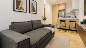 A modern living room with a grey sofa, a small side table, and a kitchen area with wooden cabinets and a stainless steel refrigerator. - w* | Amazing 2BR in the Heart of Roma Sur (Ciudad de México)