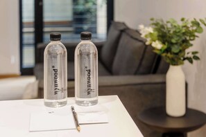 Zwei klare Wasserflaschen mit Etiketten auf einem weißen Tisch in einem modernen Zimmer mit einem grauen Sofa und einer Vase mit weißen Blumen.
