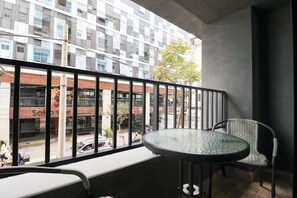 Ein Balkon mit einem runden Tisch und zwei Stühlen, mit Blick auf eine Straße mit Gebäuden und einem sichtbaren Geschäftseingang.