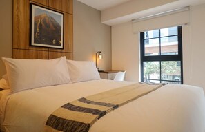 Ein Hotelzimmer mit einem großen Bett, einem Nachttisch, einer Lampe, einem Fenster mit Blick und einem gerahmten Bild an der Wand.