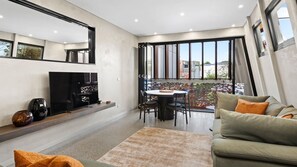 Ein modernes Wohnzimmer mit Flachbildfernseher, Esstisch mit Stühlen, einer Couch und großen Fenstern mit Blick nach draußen.