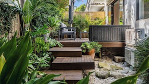 Une terrasse en bois avec des marches menant à un espace détente, entourée d’une végétation luxuriante et bordée d’un chemin en pierre.