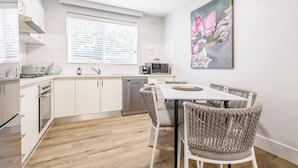 Une cuisine moderne avec des meubles blancs, une table à manger entourée de chaises en osier et un tableau représentant des fleurs accroché au mur.