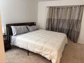 Un dormitorio con cama, mesita de noche, lámpara y ventana con cortinas.