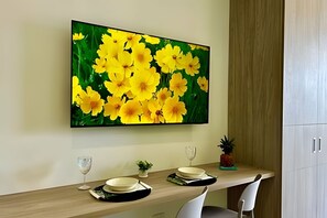 Un coin repas agrémenté d’un tableau représentant des fleurs jaunes accroché au mur, d’une table en bois, de deux chaises, d’une petite plante en pot et d’un verre à vin.