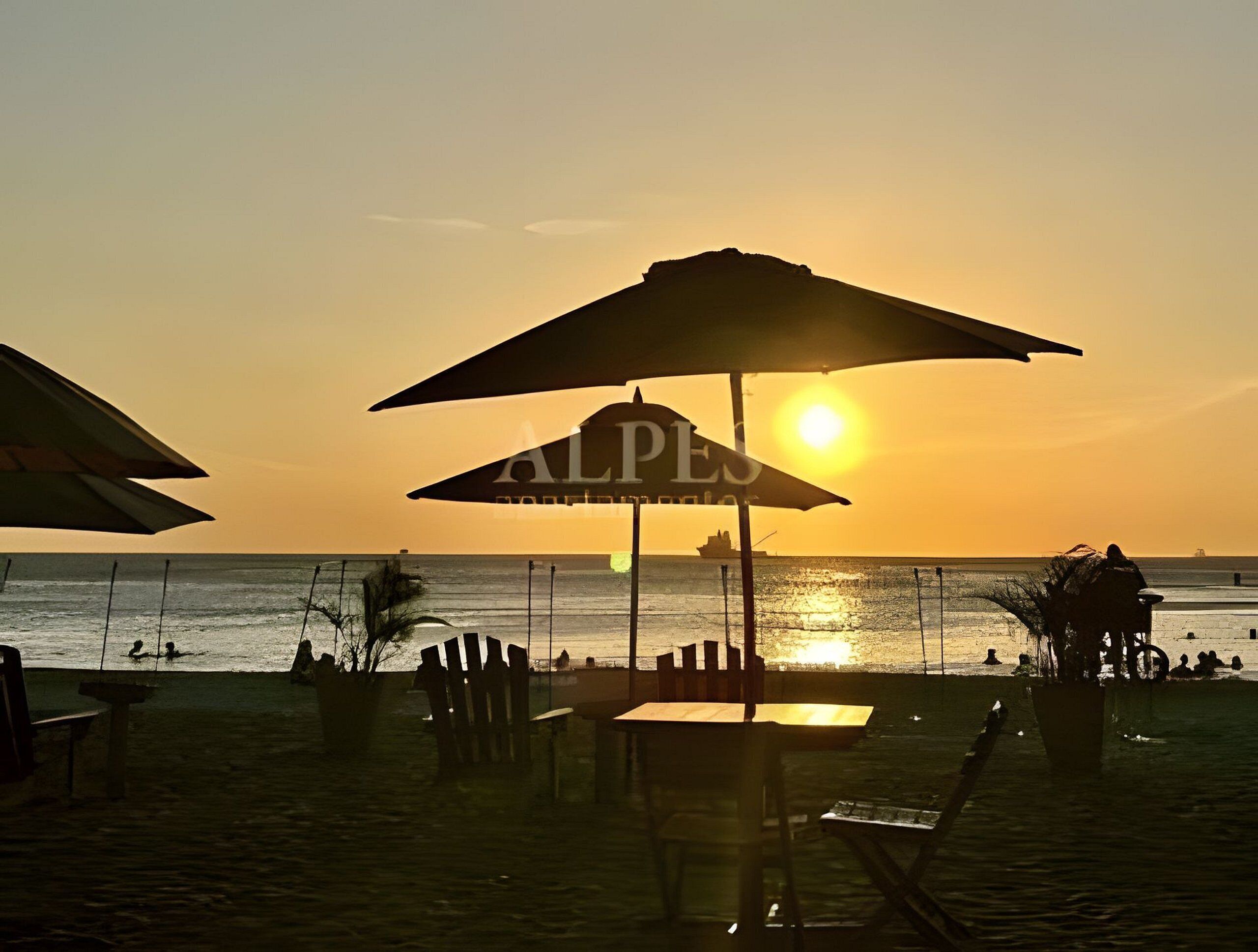 Escena de playa con sombrillas, una mesa y sillas, y un atardecer al fondo.