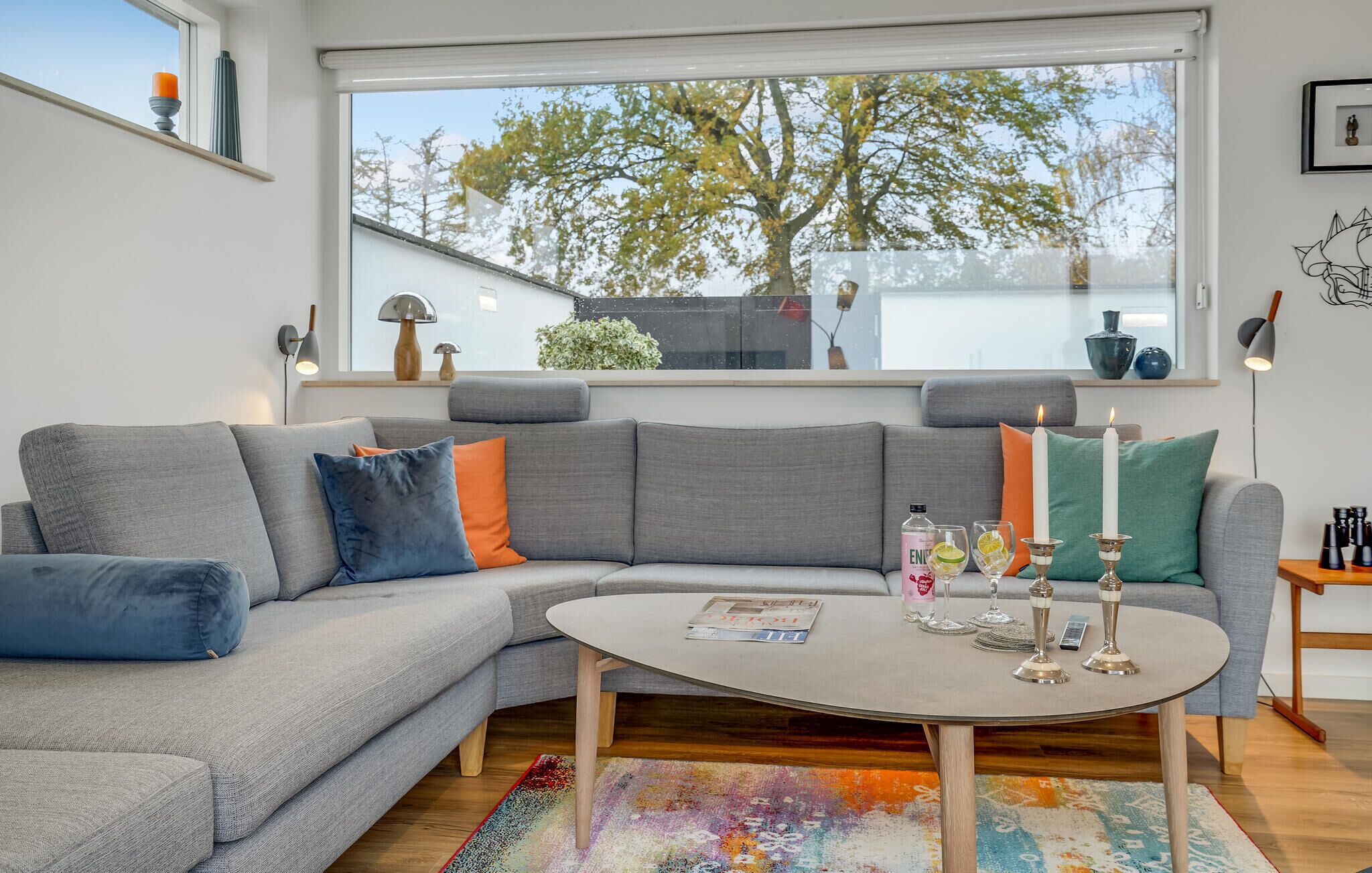 Un salon moderne avec un canapé d’angle gris, une table basse ronde et une vue sur les arbres par une grande fenêtre.