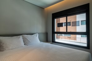 Ein Hotelzimmer mit großem Fenster, einem Bett mit weißer Bettwäsche und Blick auf ein Gebäude.