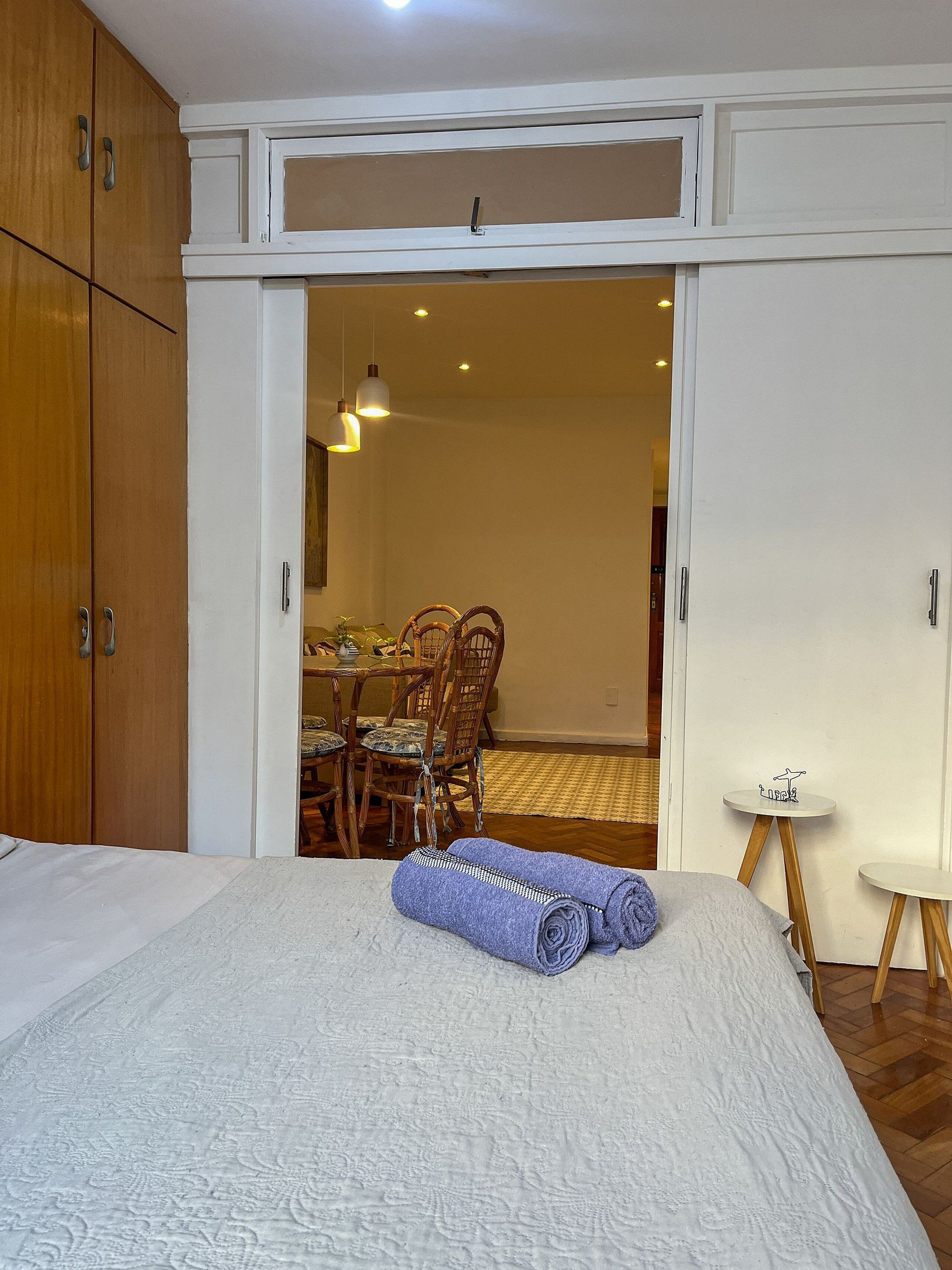 A bedroom with a bed, a rolled towel, a dining area with chairs and a table, and a small side table.