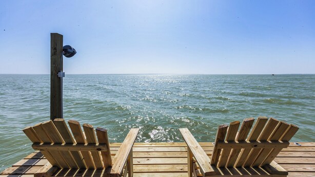 A wooden pier with two Adirondack chairs facing a calm body of water under a clear sky.