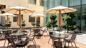 An outdoor patio with round tables and chairs, beige umbrellas, and potted plants. - BTC hotel (Malacca City)