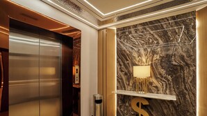 An elevator with a metallic finish, a marble wall with a geometric light fixture, and a dollar sign decoration on the floor. - BTC hotel (Malacca City)