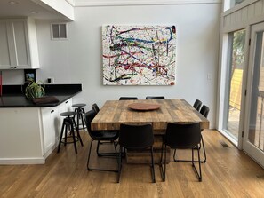 Eine moderne Küche mit Essbereich, ausgestattet mit einem Holztisch und schwarzen Stühlen, einem großen abstrakten Gemälde an der Wand und Blick durch eine Glastür.