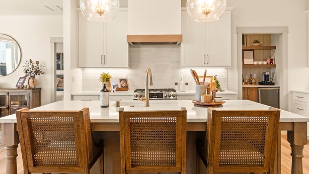 Een moderne keuken met een centraal eiland, twee hanglampen en een eethoek met drie rieten stoelen.