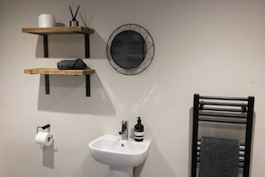 A bathroom with a white sink, a towel rack, a toilet paper holder, and two shelves with a roll of toilet paper and a bottle. - Shepherd's Hut - Sleeps 2 - Hot Tub (Northwich)