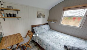 A small, single-bed room with a wooden bed frame, a small table, a chair, a shelf with storage containers, and a window with a view. - Shepherd's Hut - Sleeps 2 - Hot Tub (Northwich)
