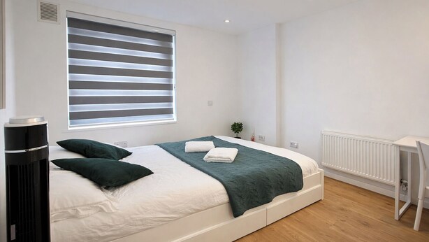 A modern bedroom with a bed, bedside table, radiator, and window with blinds. - 6 Bed House Sleeps 16 - Central London, Parking (London)