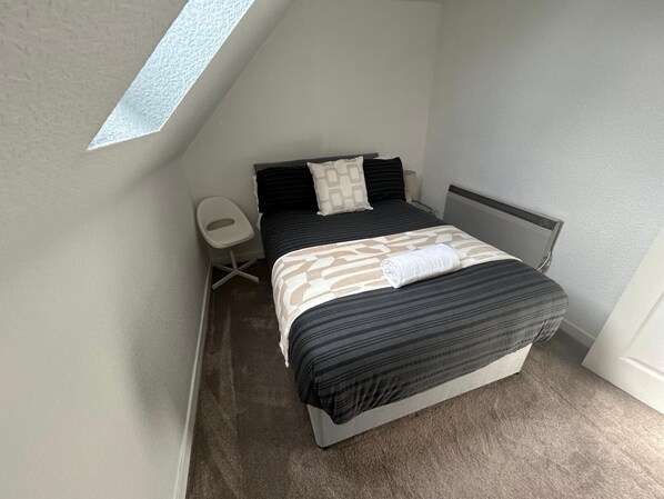 A small, modern bedroom with a sloped ceiling, a single bed, a bedside table, and a chair.