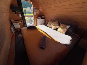Dormitorio de una cabaña de madera con cama, almohadas, una mesita y una ventana con vistas a un bosque.