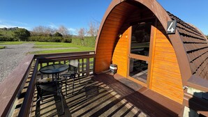 A wooden cabin with a curved roof, number 4, featuring a deck with a table and chairs, and a view of a green landscape. - Pod 4 at Flat Hills Cafe and Tourist Park - King Pod off SH1 (accessible pod) (Ōhingaiti)