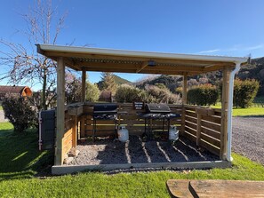 Un espace barbecue extérieur abrité, équipé d’un gril, d’une bouteille de gaz et d’un panneau indiquant qu’il faut nettoyer le barbecue après utilisation.