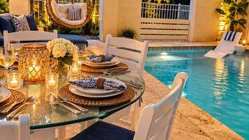 Una mesa de comedor con superficie de vidrio, sillas blancas, una piscina y una silla colgante.