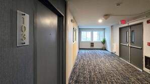 En hall med ett mattbeklätt golv, en dörr med skylten ”103” och ett fönster med en växt i.