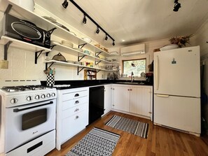 A compact kitchen with white cabinetry, a stove, a refrigerator, and a window with a view. - Fire Island Beach House (Ocean Beach)