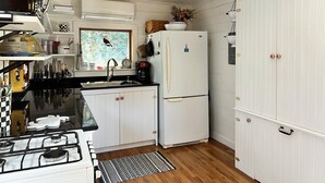 Cocina compacta con muebles blancos, estufa a gas, refrigerador y ventana con vistas.