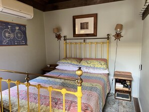 A room with a bed, a wooden headboard, a wall-mounted air conditioner, a framed picture, and a small shelf with books and headphones. - Fire Island Beach House (Ocean Beach)