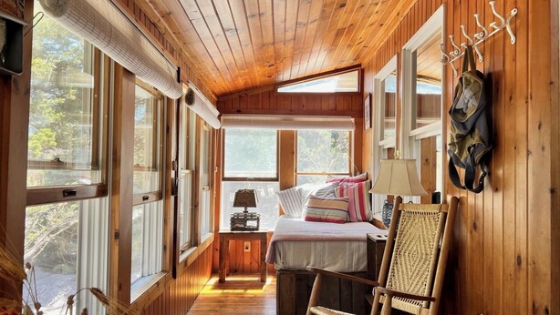 Interior de una cabaña con piso de madera, una cama con almohadas, una silla, una lámpara y un perchero en la pared.